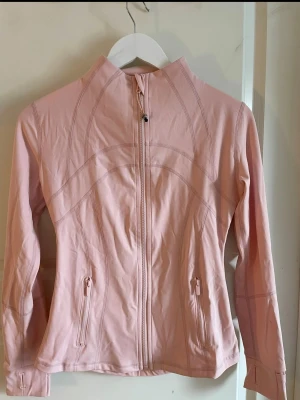 Ljusrosa träningsjacka från Lululemon - Snygg ljusrosa träningsjacka från Lululemon med figurnära passform, hel dragkedja framtill och två fickor med dragkedja. Jackan har långa ärmar och dekorativa sömmar för en sportig look. Perfekt för löpning eller gym.