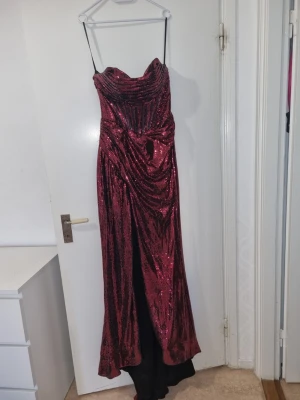 Vinröd glittrig långklänning - Säljer en vinröd långklänning med glittriga pal/fjett-detaljer och draperad framsida. Klänningen är axelbandslös och har en elegant, figurnära pass double. Perfekt för dig som vill division ut på bal eller fest. Använd en gång. Storlek 36 