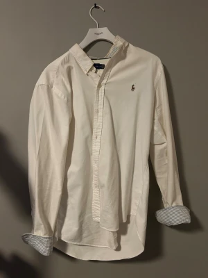 Ralph Lauren vit skjorta M – blå randig detalj - Snygg vit skjorta från Ralph Lauren. Blårandig innerkrage och i ärmslut när man viker upp dem – riktigt fin detalj.  Storlek: M Skick: bra  Perfekt både till vardag och mer uppklätt.  Finns i Göteborg. Kan skickas.