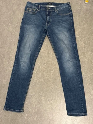 Blå slim fit jeans från Fynch-Hatton - Snygga blå slim fit jeans från Fynch-Hatton med klassisk femficksdesign och lätt slitna detaljer framtill. Jeansen har normal midja, dragkedjegylf och är tillverkade i mjuk denim med lite stretch för extra komfort. De har ett litet slitage framtill, därav priset, annars är skicket bra!