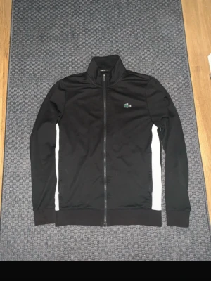 Svart Kofta från Lacoste  - Snygg svart Kofta/Jacka från Lacoste med dragkedja framtill och vita paneler längs sidorna. Klassisk passform och hög krage. Ikoniska Lacoste-loggan med krokodil på bröstet. Perfekt för sport eller chill. Materialet är mjukt och stretchigt. Tvär fet, finns inga kvar.
