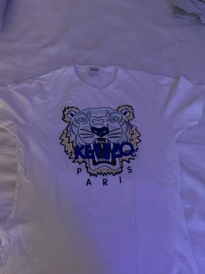 Vit Kenzo t-shirt - Säljer en vit t-shirt från Kenzo med ikoniskt tigertryck och blå text på bröstet. Klassisk passform med rund hals och korta ärmar. Perfekt statement-plagg för dig som gillar streetstyle och snygga loggor.