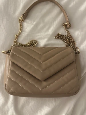 Beige quiltad handväska med kedja - Nästan helt oanvänd handväska bara utan prislapp och tar hårt i mig att sälja.Snygg beige handväska i quiltat mönster med guldfärgad kedjerem och magnetlås. Väskan har ett kuvertformat lock och innerfack med dragkedja. Perfekt accessoar för att lyfta din outfit.