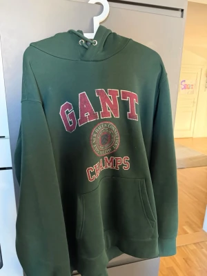 Grön hoodie från GANT med tryck - Mörkgrön hoodie från GANT med stor magficka och huva med dragsko. Framsidan har ett stort tryck i vinrött och vitt med texten 'GANT CHAMPS' och ett emblem. Perfekt för en avslappnad och sportig stil. Använd fåtal gånger.