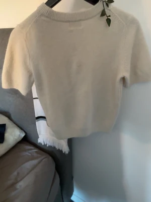 Beige stickad kortärmad tröja - Snygg beige stickad tröja med korta ärmar och rund halsringning. Tröjan har en enkel och stilren design med ribbstickade detaljer vid halsen. Perfekt att matcha med jeans eller kjol för en avslappnad look.