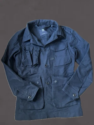 Svart utilityjacka från G star RAW - Stilren svart utilityjacka från G star RAW med flera praktiska fickor framtill och klassisk krage. Jackan har knäppning med knappar och långa ärmar. Perfekt för dig som gillar funktionell och snygg design.