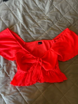 Topp från Gina Tricot - Trendig röd offshoulder topp från Gina Tricot med volangkant och knyt framtill. Toppen har korta puffärmar och är tillverkad i ett lätt, strukturerat material som passar perfekt till sommarens outfits. Storlek XS, inte kommit till användning. Så är helt ny.