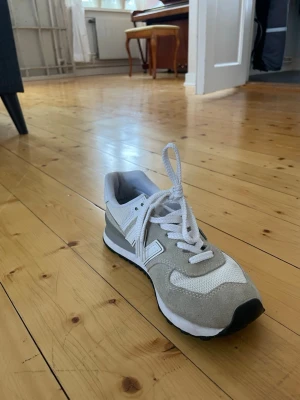 New Balance grå/vita sneakers - Snygga New Balance sneakers i grått och vitt med klassisk N-logga på sidan. Skorna har mesh och mocka-material, vita snören och svart yttersula. Perfekt för dig som gillar en clean och sportig look.