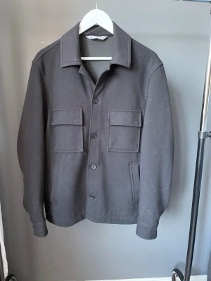 Svart overshirt från Zara - Stilren svart overshirt från Zara med klassisk krage och fyra fickor framtill, varav två med lock. Jackan har raka ärmar och stängs med knappar. Perfekt för lager-på-lager och enkel att matcha med olika outfits.