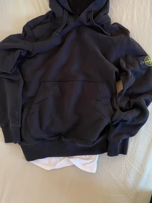 Svart hoodie från Stone Islands - Snygg svart hoodie från Stone Island med klassisk huva och känguruficka framtill. Ikonisk logotyp-patch på ärmen och mjukt material som känns skönt mot huden. Perfekt för en avslappnad streetwear-look.