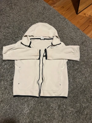 Beige hoodie från Nike med svarta detaljer - Snygg beige hoodie från Nike med svarta detaljer och dragkedja framtill. Hoodien har en huva, svarta kantband och en extra ficka med dragkedja på ärmen. Perfekt för dig som gillar sportig och clean stil.