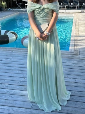 By Malina Maxiklänning Selma Dress, Small - Säljer en elegant ljusgrön långklänning med offshoulder och draperad överdel från By Malina. Klänningen har en markerad midja, slits framtill och ett flytande, luftigt tyg som ger en romantisk känsla. Perfekt för bal eller student. Använd en dag, men lite smutsig längst ner, se foto. För att hålla fast offshoukder, är ett fint band sytt i ryggen Nypris 2700:-. 