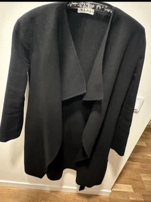 Svart kappa från Wera Stockholm - Stilren svart kappa från Wera Stockholm med öppen front och sjalkrage. Kappan har ett avtagbart midjebälte och raka ärmar. superfin ullkappa i stl 36, passar dock mig som är 175 cm och väger drygt 75. Stor i storleken med andra andra ord. Använd Max 5 gånger. Det enda anmärkningsbara är att sömmen har släppt i nacken på två ställen se bilder, ena sömmen har jag sytt för hand. Kan sys om man har symaskin eller för hand om man är händig. I övrigt inget att anmärka på. Nypris 2500 kr.   