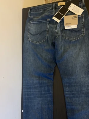 Slim Glenn blå jeans från Jack & Jones - Säljer ett par Slim Glenn jeans från Jack & Jones i klassisk blå denim. Jeansen har slim fit passform, fem fickor och snygga slitningar framtill. Materialet är mjuk bomullsdenim med stretch för extra komfort. Perfekta för en avslappnad och trendig look.