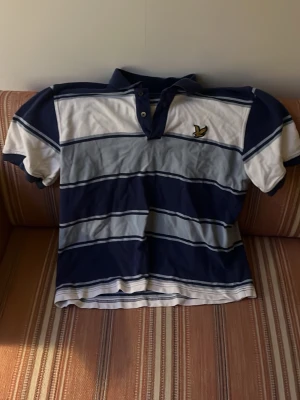 Randig pikétröja från Lyle & Scott - Snygg pikétröja från Lyle & Scott med breda horisontella ränder i vitt, blått och grått. Tröjan har korta ärmar, krage med knappar och den klassiska loggan broderad på bröstet. Perfekt för en avslappnad stil. 