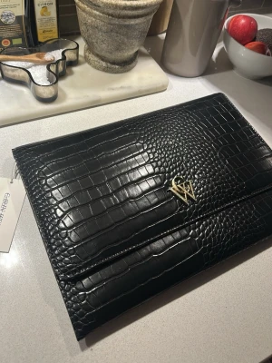 Svart kuvertväska i krokopräglat skinn CW - Stilren svart kuvertväska från Wera i krokopräglat skinn med guldfärgad logga framtill. Väskan har magnetlås och ett mönstrat innerfoder med Wera-loggor. Perfekt för att bära det viktigaste till utekvällen eller eventet.