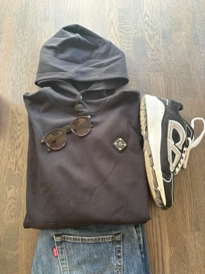 Svart hoodie från j.lindeberg - Säljer en svart hoodie från J.Lindeberg med knappdetaljer vid halsen istället för snören och en liten patch på bröstet. Hoodien är i fint och välanvänd skick, frågor eller fler bilder? - skriv till mig.