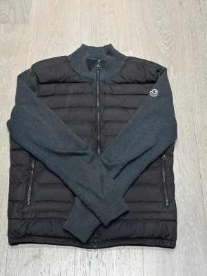 Moncler grå cardiganjacka - Snygg cardiganjacka från Moncler med quiltad svart front och grå stickade ärmar. Jackan har dragkedja framtill, ribbad krage och muddar samt Moncler-logga på ärmen. Perfekt för lager-på-lager och stilren look.