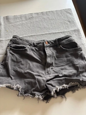 Gråa jeansshorts från H&M Divided - Snygga gråa jeansshorts från H&M Divided med fransig och rå kant nertill. Klassisk femficksmodell med knapp och dragkedja framtill. Perfekta för en avslappnad stil under varma dagar.