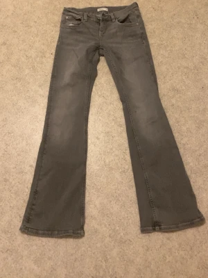 Grå bootcut jeans med fickdetaljer - Snygga grå bootcut jeans med klassiska fem fickor och dekorativa sömmar på bakfickorna. Jeansen har en lätt utsvängd passform nertill och är tillverkade i denim med stretch för extra komfort. Perfekta för en avslappnad och trendig look.