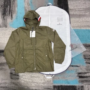 Olivgrön vindjacka från Moncler - Snygg olivgrön vindjacka från Moncler med huva och dragkedja framtill. Jackan har justerbara snören vid huvan, Moncler-logga på ärmen och detaljer i rött, vitt och blått vid huvkanten. Tillverkad i lätt, vattenavvisande material. Perfekt för blåsiga dagar. Storlek 3 passar M och L. Kan gå ner i pris vid snabb affär. Hör av dig vid funderingar! :)