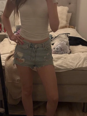 Ljusblå denimshorts med slitningar - Säljer ett par ljusblå denimshorts med råa kanter och slitna detaljer. Shortsen har fem fickor, knappgylf och en klassisk passform som sitter midwaist. Perfekta för sommaren och ger en avslappnad vibe. Ingen aning vart dem är köpte för jag har fått dem av en kompis. Men skulle gissa på new yorker eller hm. 