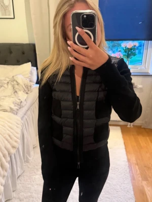 Moncler cardigan  - Snygg svart cardiganjacka från Moncler med quiltad front och fyra fickor. Jackan har dragkedja, ribbade muddar och Moncler-logga på ärmen. Materialmix med vadderad front och stickade ärmar ger en modern look.