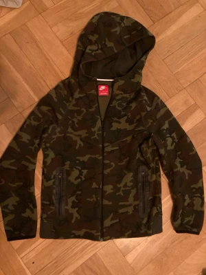 Nike camo hoodie med dragkedja, storlek junior L - Snygg hoodie från Nike i camomönster med dragkedja framtill och två fickor med dragkedja. Huva med skön passform och mjukt material. Perfekt för dig som gillar streetwear och vill sticka ut lite extra.