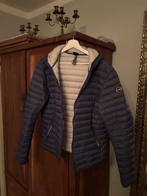 Dunjacka från Colmar  - Snygg blå dunjacka från Colmar med quiltad design och huva. Jackan har dragkedja framtill, två sidofickor med dragkedja och ljusgrått foder. Perfekt för kalla vinterdagar och riktigt skön att bära. Storlek 52 vilket motsvarar M/L. Nästintill ny! Använd ett fåtal gånger!