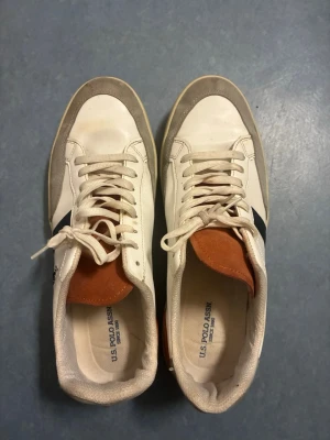 Vita sneakers från U.S. Polo Assn. - Snygga vita sneakers från U.S. Polo Assn. med grå mockadetaljer, svart sidostrip och brun mocka på plösen. Klassisk logga på sidan och vita snören. Ovandel i läder och mocka, perfekt för en stilren look.