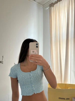 Ljusblå blommig croppad topp - Superfin ljusblå croppad topp med små vita blommor och korta ärmar. Toppen har knappar framtill och en söt, lätt vågig kant nertill. I väldigt bra skick! Skriv för fler bilder