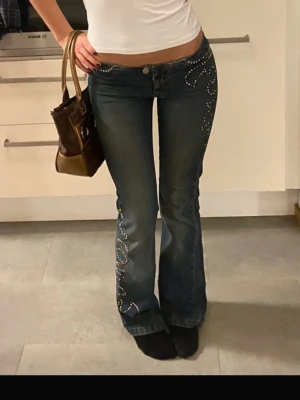 Bootcut jeans med strassdetaljer - Mörkblå bootcut jeans med färgglada strassdetaljer längs fickor och ben. Jeansen har låg midja och utsvängda ben, vilket ger en snygg 2000-talsvibe. Perfekta för dig som gillar coola detaljer