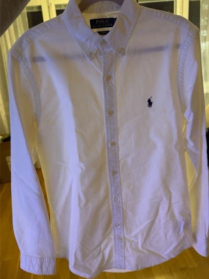 Vit skjorta från Polo Ralph Lauren - Klassisk vit skjorta från Polo Ralph Lauren med lång ärm och knappar framtill. Skjortan har en broderad marinblå logga på bröstet och är tillverkad i bomull. Perfekt för en stilren och tidlös look. Den är knappt använd. Sälj pga den är för liten