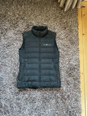 Svart dunväst från Salomon - Snygg svart dunväst från Salomon med hög krage och två dragkedjeförsedda fickor. Västen har quiltad design och är tillverkad i lätt, värmande material med 800 fill power dun. Perfekt för lager-på-lager och outdoor-vibes.