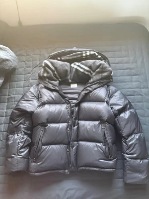 Svart buberry pufferjacka  - Snygg svart pufferjacka med stor vaddering och dragkedjor på fickorna. Jackan har en avtagbar huva med grått och vitt randigt innerfoder. Perfekt för kalla dagar och riktigt trendig look.