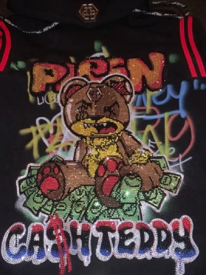 Philipp Plein hoodie M - Svart hoodie från Philipp Plein med huva och dragsko. Framsidan har en stor, glittrig björn med kedjor och pengar, samt graffiti-inspirerad text och detaljer i rött, gult och grönt. Röda ränder på axlarna och tydlig streetwear-vibe.