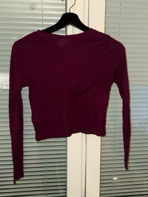 Mörklila ribbad croppad topp - En mörklila  topp med lång ärm och ribbad struktur. Toppen har en rund halsringning och är figurnära i modellen. Perfekt att styla med höga jeans eller kjol för en trendig look.
