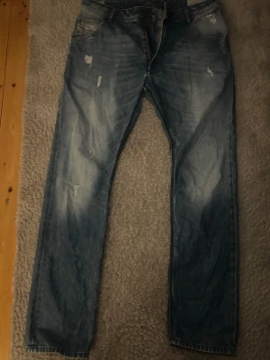 Diesel - Säljer ett par blå diesel jeans med raka ben och snygga slitningar framtill. Jeansen har klassisk femficksdesign och ljus tvätt med slitna detaljer för en cool look. Perfekt för dig som gillar en avslappnad stil.