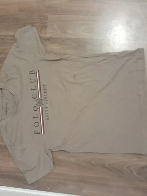 Beige t-shirt Polo Club Saint Vincent - Snygg beige t-shirt från Polo Club Saint Vincent med tryck framtill och klassisk passform. T-shirten har korta ärmar och rund halsringning. Perfekt för en avslappnad och stilren look.Priser kan diskutera!