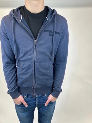 Blå zip hoodie från Woolrich - Mörkblå hoodie från Woolrich med dragkedja framtill och broderad logga 'WOOLRICH 1830' på bröstet. Tröjan har huva med dragsko, två stora fickor fram och ribbade muddar vid ärmslut och nederkant. Tillverkad i mjuk bomullsblandning, perfekt för en avslappnad stil.