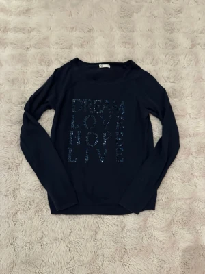 Mörkblå långärmad topp med strass - Mörkblå långärmad topp från Cubus med texten 'DREAM LOVE HOPE LIVE' framtill i glittriga strasstenar. Rund halsringning och mjukt material som känns skönt mot huden. Perfekt för dig som gillar statement-plagg med lite extra bling. 