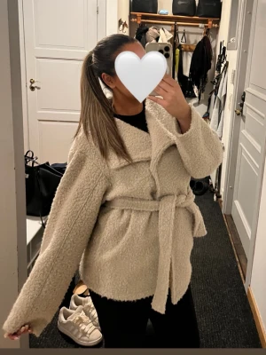 Beige teddyjacka/kappa - Mysig och trendig beige teddyjacka med knytskärp i midjan. Jackan har en bred krage och långa ärmar, perfekt för att hålla värmen. Materialet är fluffigt och mjukt, vilket ger en cool och avslappnad look.
