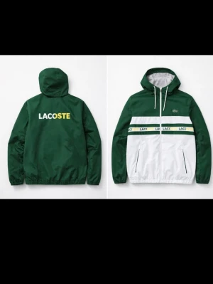 Grön och vit vindjacka från Lacoste - Snygg vindjacka från Lacoste i grönt och vitt med dragkedja framtill och huva. Jackan har vita partier på framsidan, gröna ärmar och rygg samt en rand med Lacoste-loggan i grönt och gult över bröstet. Två fickor med dragkedja och justerbar huva med snören.