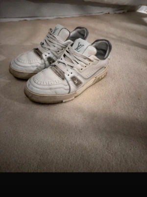 Louis Vuitton vita sneakers - Snygga vita Louis Vuitton sneakers med grå detaljer och klassisk LV-logga på sidan. Skorna har perforerade partier, tjock sula och är gjorda i läder. Perfekta för dig som vill ha en exklusiv och trendig look.