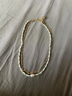 Tvådelat halsband med pärlor och guldkedja - Säljer ett snyggt tvådelat halsband med ljusblå och vita pärlor samt små guldfärgade detaljer. Ena delen är en tunn guldkedja och den andra består av pärlor med en större vit pärla i mitten. Perfekt för att ge din outfit en trendig touch.