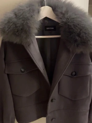 Grå Meotine jacka  - Stilren grå kappa från Meotine med. Kappan har två stora bröstfickor med lock och knappar framtill. Kortare modell som ger en modern siluett. Perfekt för dig som vill ha en trendig och varm jacka i vår. XS/S