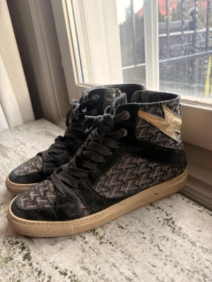 Zadig & Voltaire svarta high top sneakers - Snygga high top sneakers från Zadig & Voltaire med svart och grått mönster, svarta mockadetaljer och en guldig blixt på sidan. Skorna har tjocka svarta snören och beige sula. Perfekta för dig som vill sticka ut med en cool streetstyle.