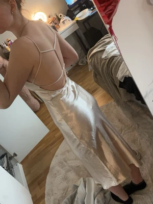 Champagnefärgad långklänning  - Säljer en elegant långklänning i champagnefärgad satin från ginatricot. Klänningen har tunna axelband, öppen rygg med snygga banddetaljer och en mjukt skimrande yta. Perfekt för dig som vill ha en stilren och trendig look. Den är inte använd men den har lite gaderobsskador då den legat i gaderoben länge.