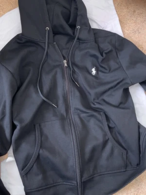 Svart zip hoodie från Polo Ralph Lauren - Säljer en svart hoodie med dragkedja från Polo Ralph Lauren. Klassisk design med vit broderad logga på bröstet, huva med dragsko och två stora fickor framtill. Tillverkad i mjukt bomullsmaterial som är skönt att ha på sig.