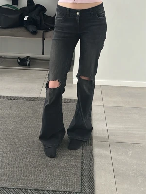 Svarta slitna bootcut jeans - Säljer ett par svarta bootcut jeans med stora slitningar över knäna. Jeansen har normal midja på mig men räknas som låg egentligen , fem fickor och råa kanter vid hålen. Perfekta för en avslappnad och trendig look. Materialet är klassisk denim i bomull.
